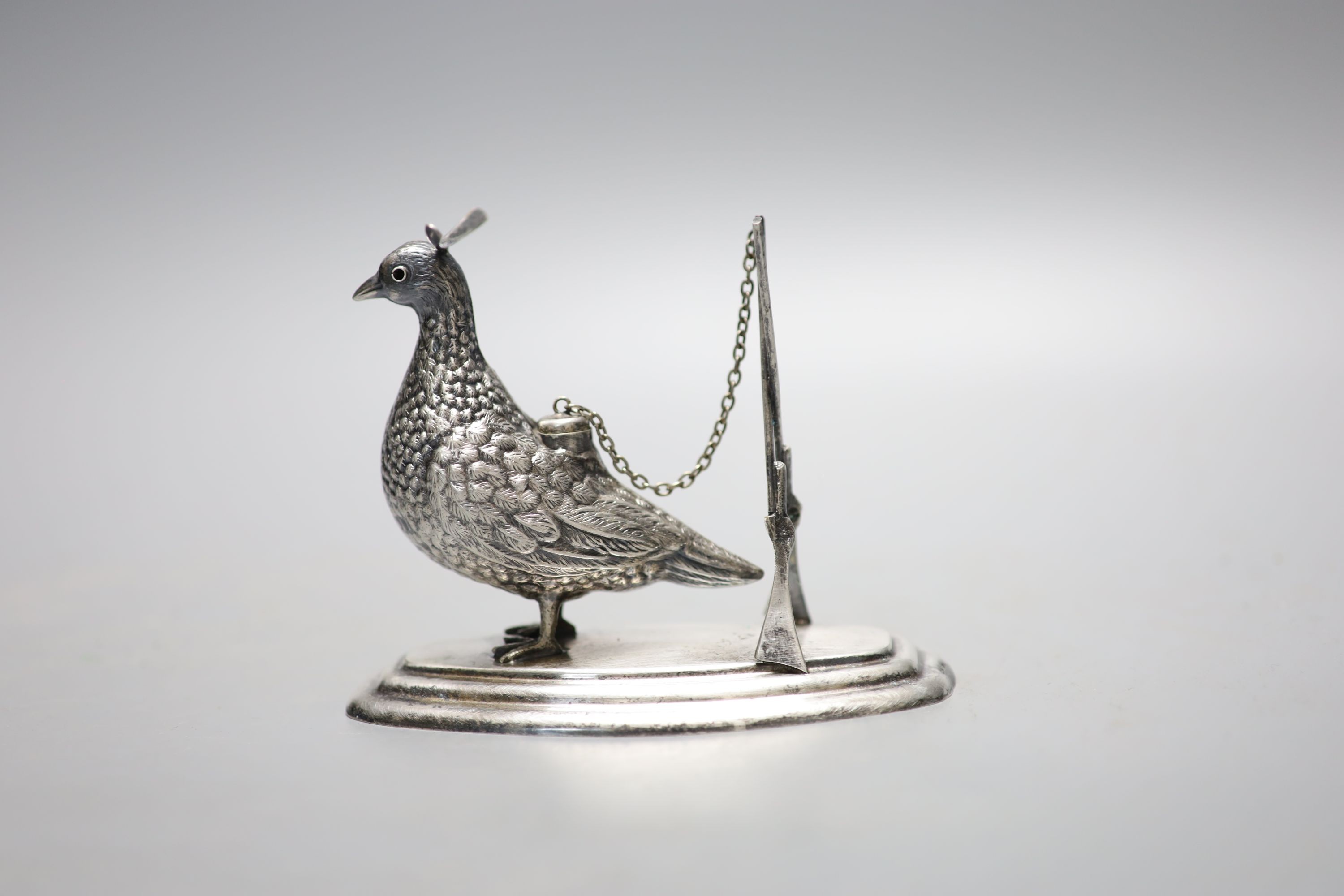 A continental novelty white metal brand balloon warmer, modelled and a game bird with crossed rifles, on a shaped oval base, 13.1cm, indistinct stamp.